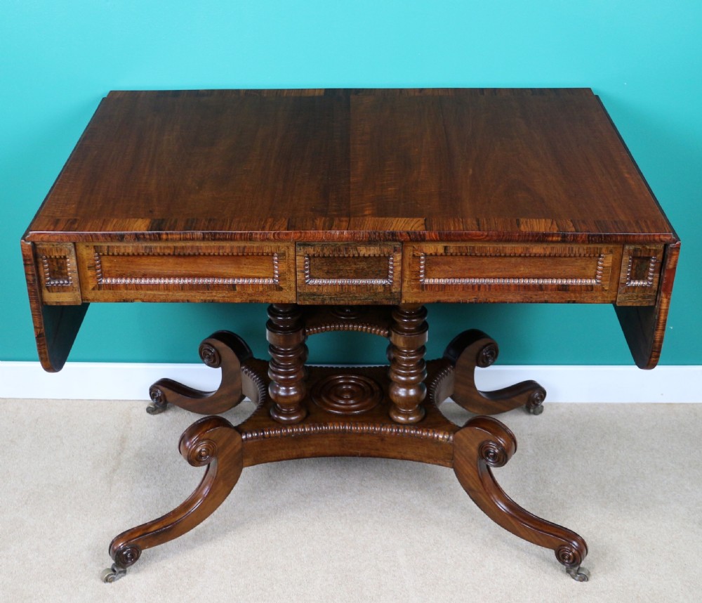 A Regency mahogany and rosewood crossbanded sofa table featuring drop leaves, bead-moulded frieze drawers, turned support pillars, and splayed legs on brass castors. View X