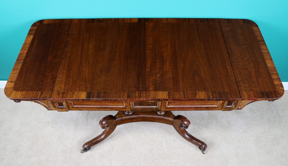 A Regency mahogany and rosewood crossbanded sofa table featuring drop leaves, bead-moulded frieze drawers, turned support pillars, and splayed legs on brass castors. View XI