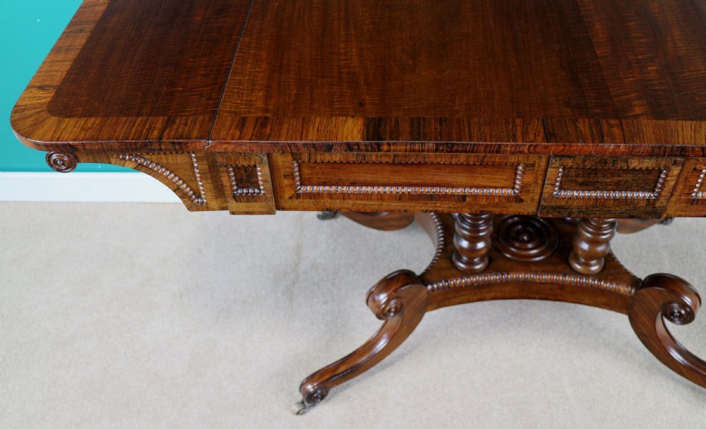 A Regency mahogany and rosewood crossbanded sofa table featuring drop leaves, bead-moulded frieze drawers, turned support pillars, and splayed legs on brass castors. View XII