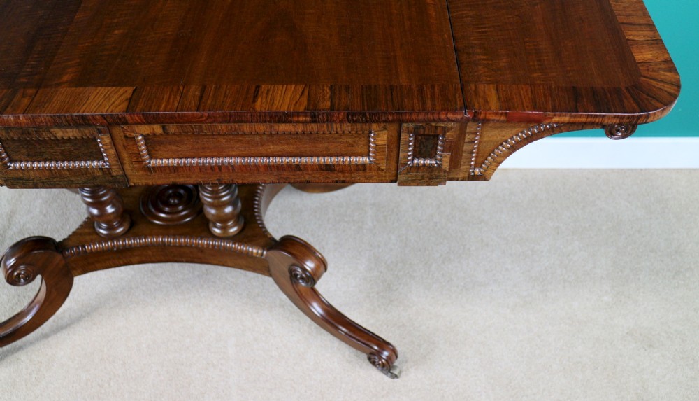 A Regency mahogany and rosewood crossbanded sofa table featuring drop leaves, bead-moulded frieze drawers, turned support pillars, and splayed legs on brass castors. View XIII
