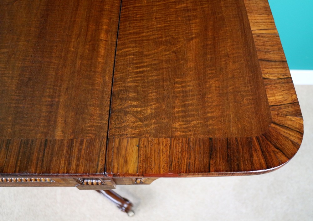 A Regency mahogany and rosewood crossbanded sofa table featuring drop leaves, bead-moulded frieze drawers, turned support pillars, and splayed legs on brass castors. View XIV