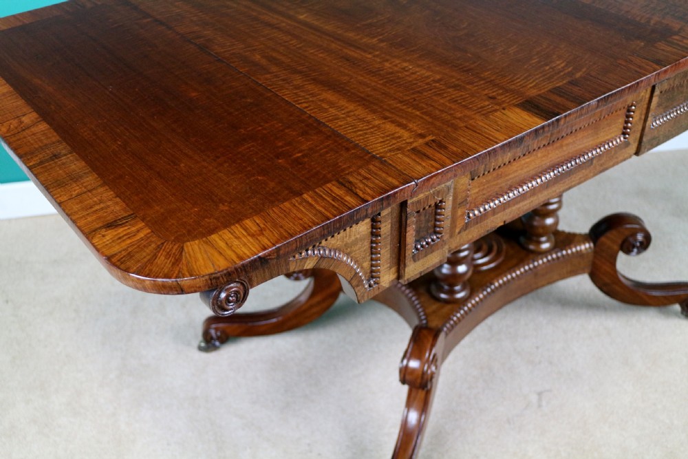 A Regency mahogany and rosewood crossbanded sofa table featuring drop leaves, bead-moulded frieze drawers, turned support pillars, and splayed legs on brass castors. View XV