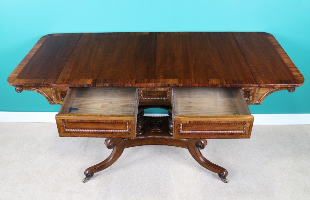 A Regency mahogany and rosewood crossbanded sofa table featuring drop leaves, bead-moulded frieze drawers, turned support pillars, and splayed legs on brass castors. View XVI