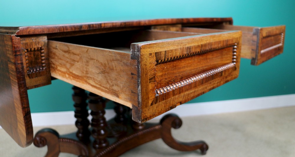 A Regency mahogany and rosewood crossbanded sofa table featuring drop leaves, bead-moulded frieze drawers, turned support pillars, and splayed legs on brass castors. View XVII