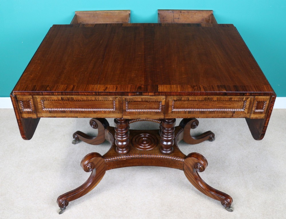 A Regency mahogany and rosewood crossbanded sofa table featuring drop leaves, bead-moulded frieze drawers, turned support pillars, and splayed legs on brass castors. View XIX