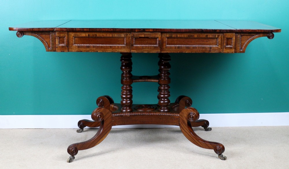 A Regency mahogany and rosewood crossbanded sofa table featuring drop leaves, bead-moulded frieze drawers, turned support pillars, and splayed legs on brass castors. View XX