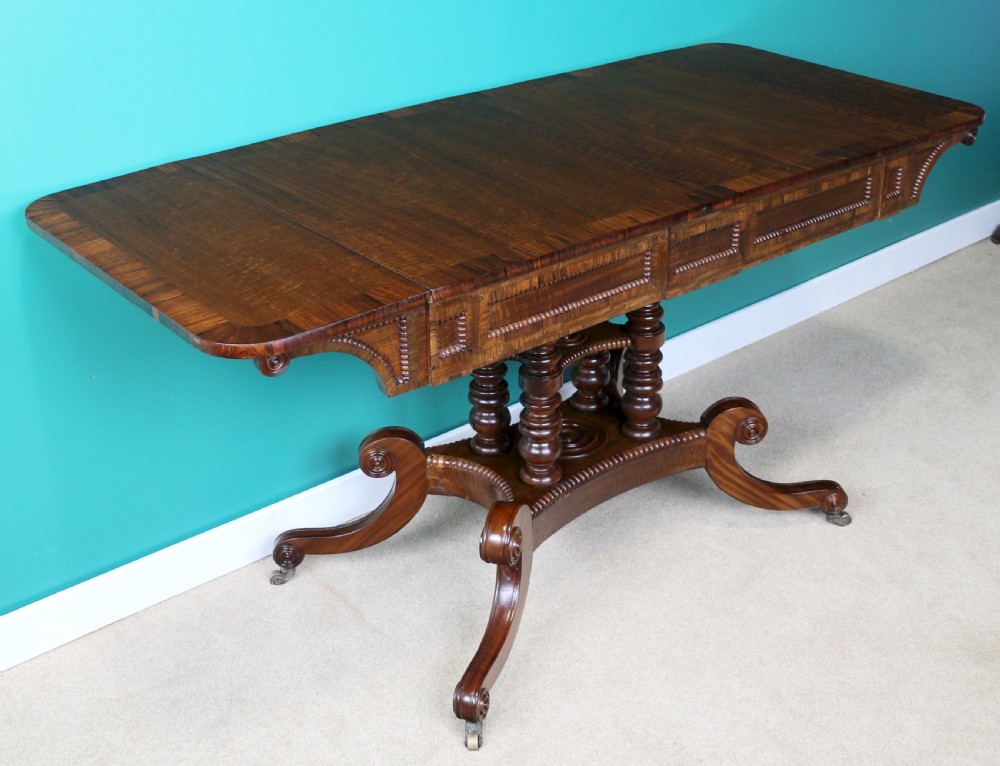 A Regency mahogany and rosewood crossbanded sofa table featuring drop leaves, bead-moulded frieze drawers, turned support pillars, and splayed legs on brass castors. View XXI