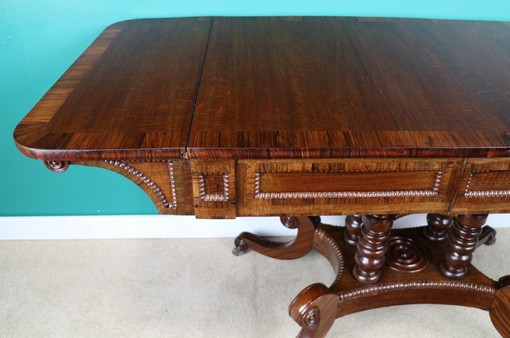 A Regency mahogany and rosewood crossbanded sofa table featuring drop leaves, bead-moulded frieze drawers, turned support pillars, and splayed legs on brass castors. View XXII