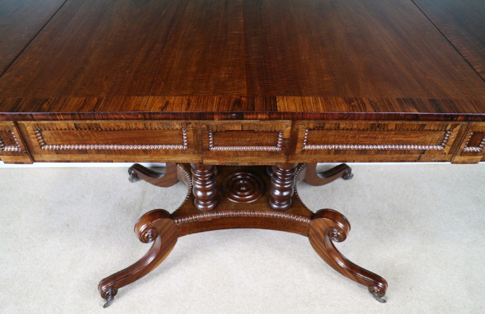A Regency mahogany and rosewood crossbanded sofa table featuring drop leaves, bead-moulded frieze drawers, turned support pillars, and splayed legs on brass castors. View XXIII