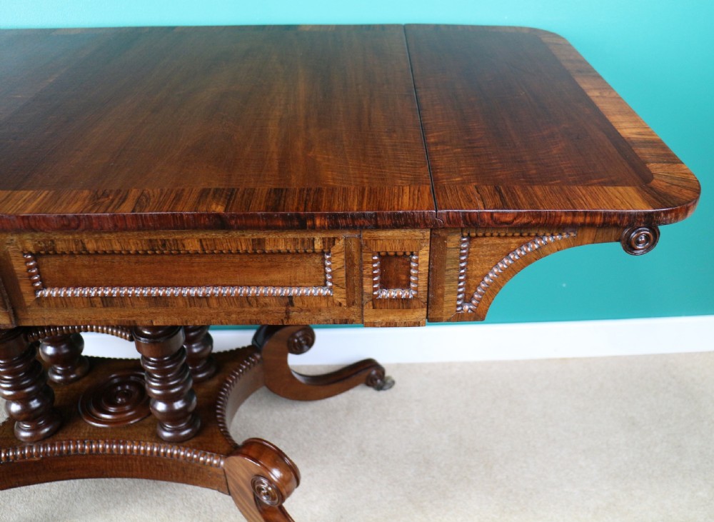 A Regency mahogany and rosewood crossbanded sofa table featuring drop leaves, bead-moulded frieze drawers, turned support pillars, and splayed legs on brass castors. View XXIV