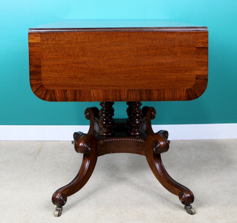 A Regency mahogany and rosewood crossbanded sofa table featuring drop leaves, bead-moulded frieze drawers, turned support pillars, and splayed legs on brass castors. View XXVIII