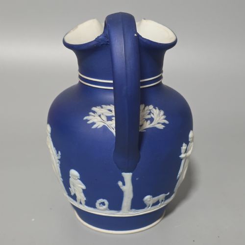 fig 1. A cobalt blue stoneware jug featuring white raised neoclassical figures in relief with a high arched handle and curved spout.
