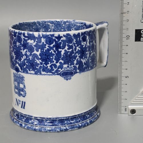 fig 5. A cylindrical blue and white transferware ceramic tankard featuring a floral upper border and a coat of arms crest with the text No 11.