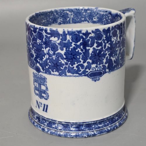 fig 6. A cylindrical blue and white transferware ceramic tankard featuring a floral upper border and a coat of arms crest with the text No 11.