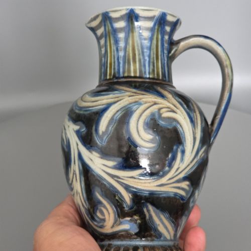 fig 1. A Victorian salt-glazed stoneware jug decorated with incised blue and cream foliage patterns, a ribbed neck, and a loop handle against a dark brown ground.