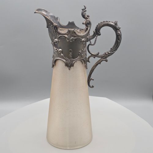 fig 1. An antique French Rococo style silver-plated claret jug featuring a tapered ribbed clear glass body and an ornate scrolling silver spout and handle.