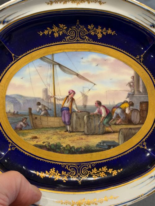 fig 2. An oval French Sevres porcelain dish featuring a deep cobalt blue rim decorated with intricate gold leaf gilding surrounding a central hand-painted maritime harbour scene.