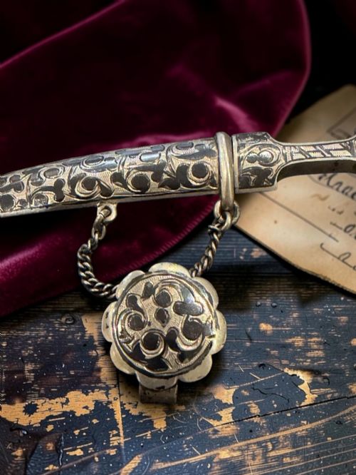 fig 1. An antique Caucasian curved silver miniature dagger brooch featuring black niello scrollwork motifs and an attached suspension chain with a matching circular clasp.