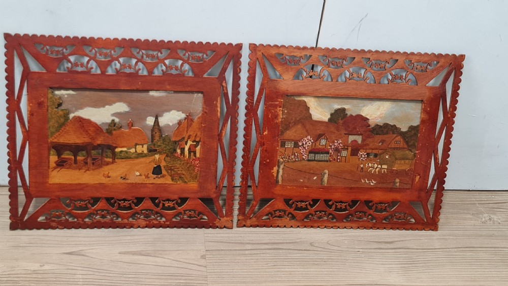 A pair of framed three-dimensional wooden folk art pictures featuring rural village scenes set within fretted reddish-brown frames.