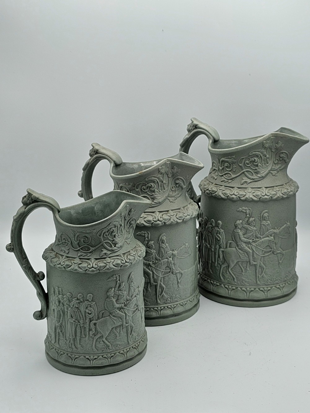 A set of three graduated sage green ceramic jugs decorated with detailed relief-moulded scenes of knights and foliate scrollwork.