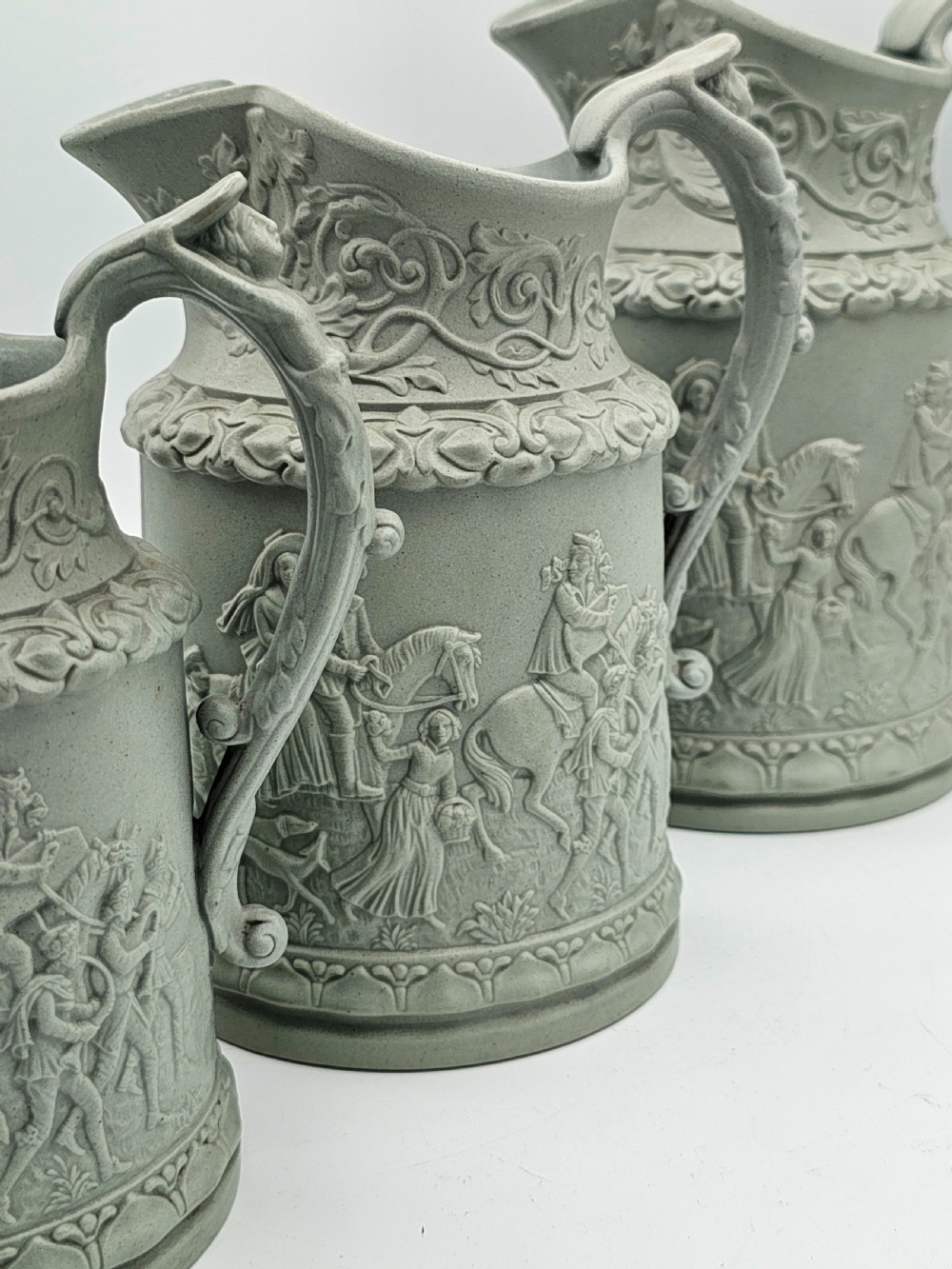 A set of three graduated sage green ceramic jugs decorated with detailed relief-moulded scenes of knights and foliate scrollwork. View V