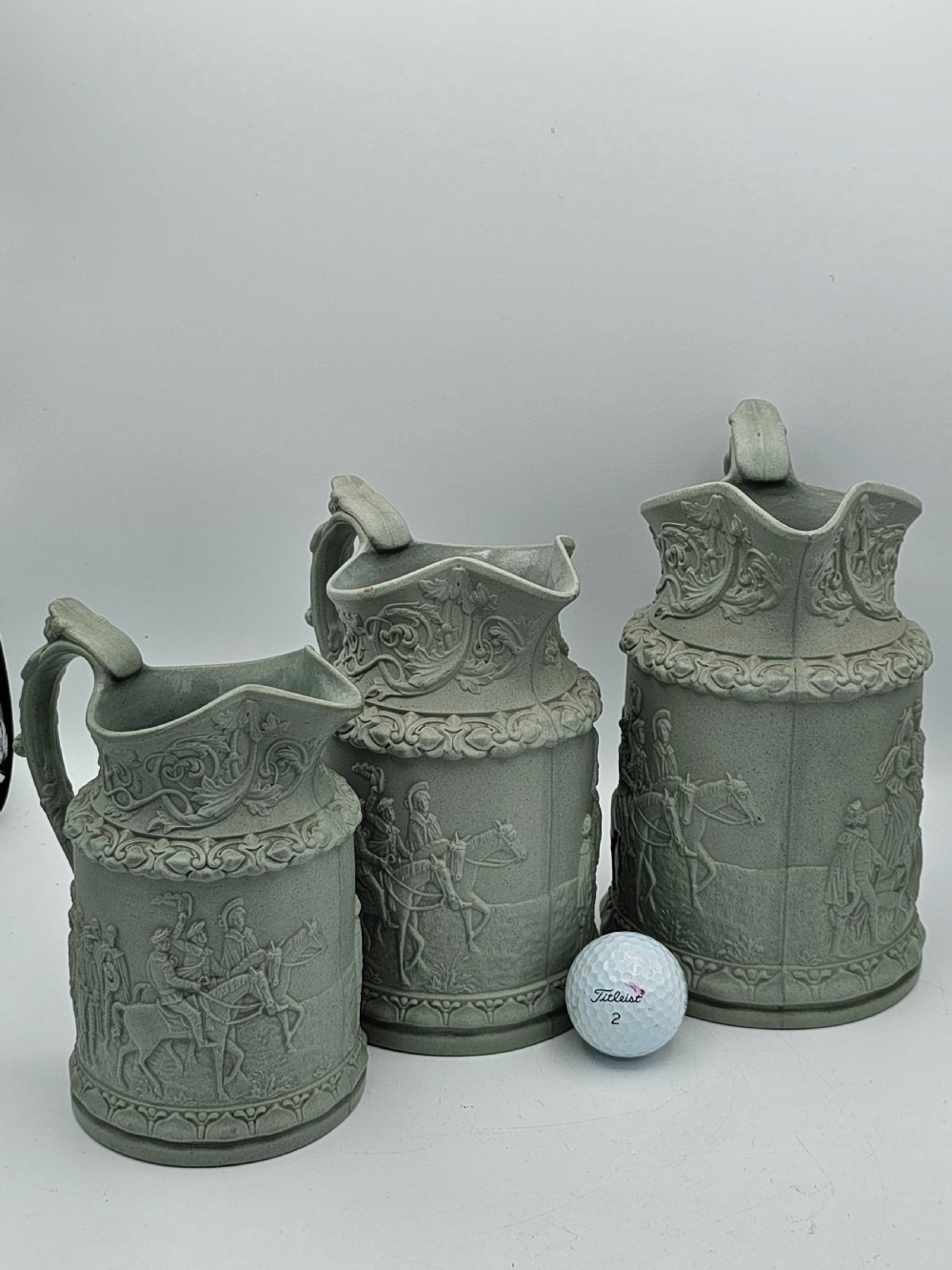 A set of three graduated sage green ceramic jugs decorated with detailed relief-moulded scenes of knights and foliate scrollwork. View IX