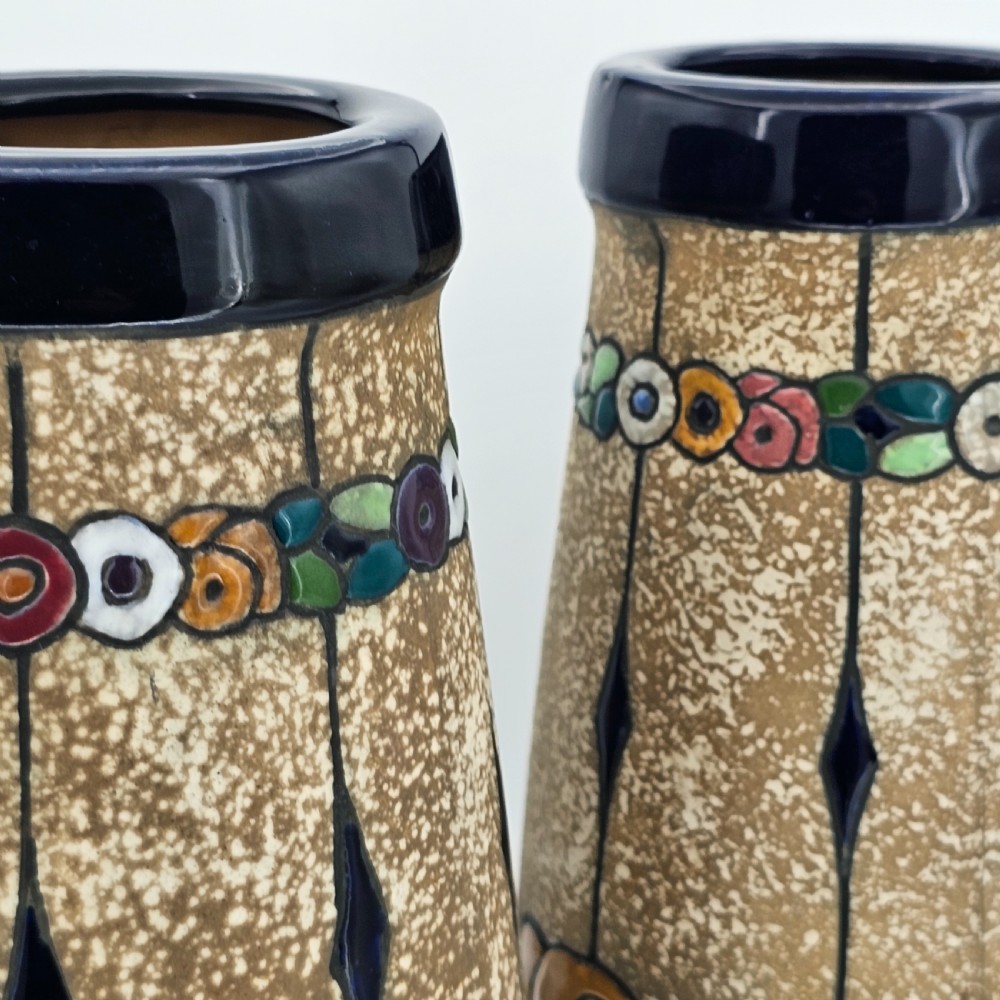 A pair of cylindrical Austrian ceramic vases featuring a textured beige ground, black rim, vertical cutouts, and colourful enamelled floral bands. View X