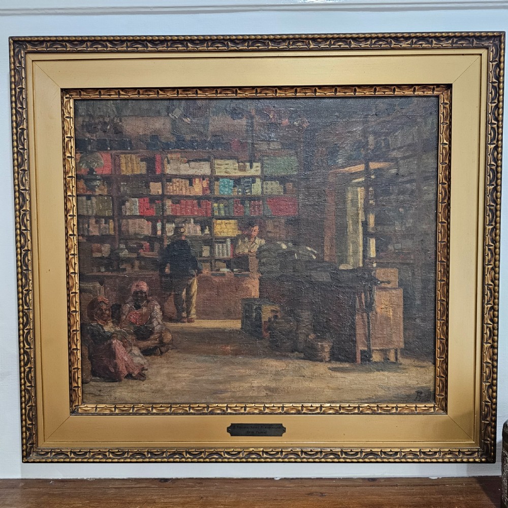 A framed oil painting depicting the interior of a village store in Namaqualand with seated figures and shelves of merchandise, set within a wide gold moulded frame.