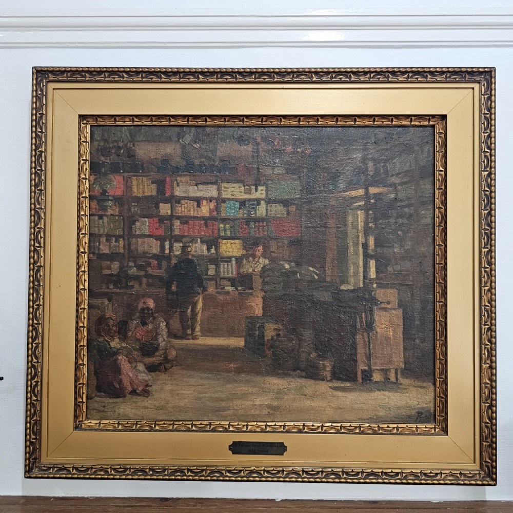 A framed oil painting depicting the interior of a village store in Namaqualand with seated figures and shelves of merchandise, set within a wide gold moulded frame. View V