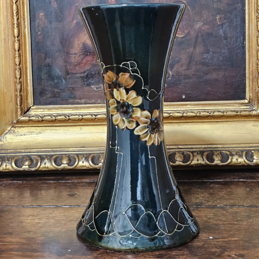 A dark green glazed earthenware vase with a flared top and base, featuring tube-lined ochre floral motifs and geometric line patterns.