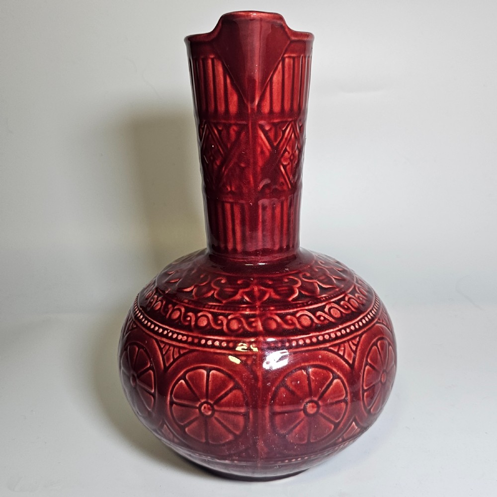 A dark red glazed earthenware jug featuring relief-moulded geometric patterns, a bulbous lower section, a fluted neck, and a curved handle. View II
