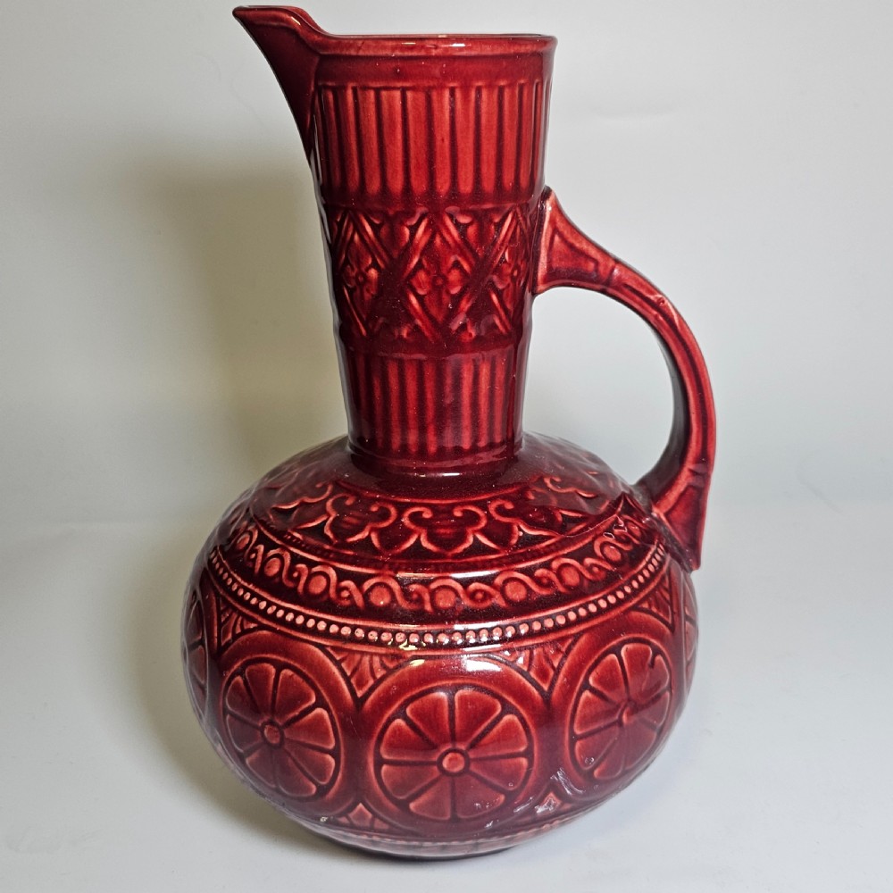 A dark red glazed earthenware jug featuring relief-moulded geometric patterns, a bulbous lower section, a fluted neck, and a curved handle.