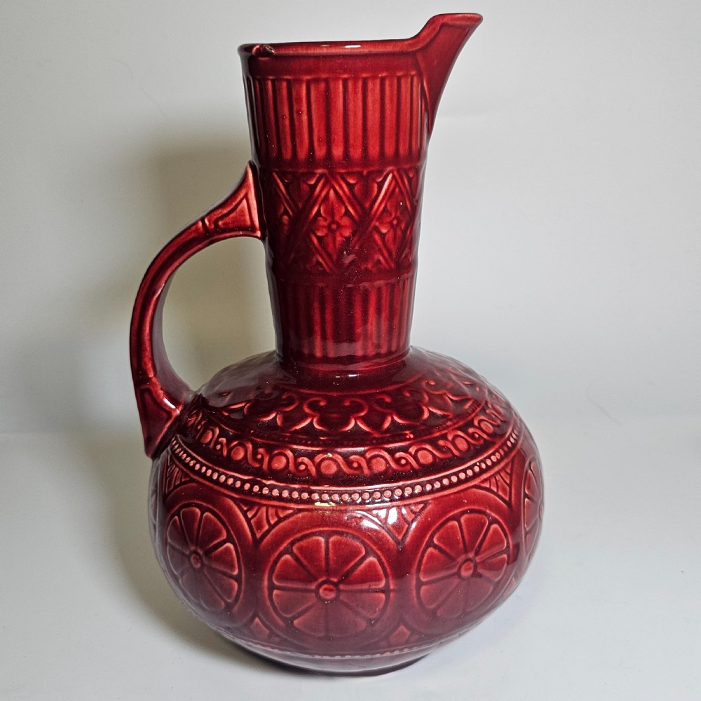 A dark red glazed earthenware jug featuring relief-moulded geometric patterns, a bulbous lower section, a fluted neck, and a curved handle. View VI