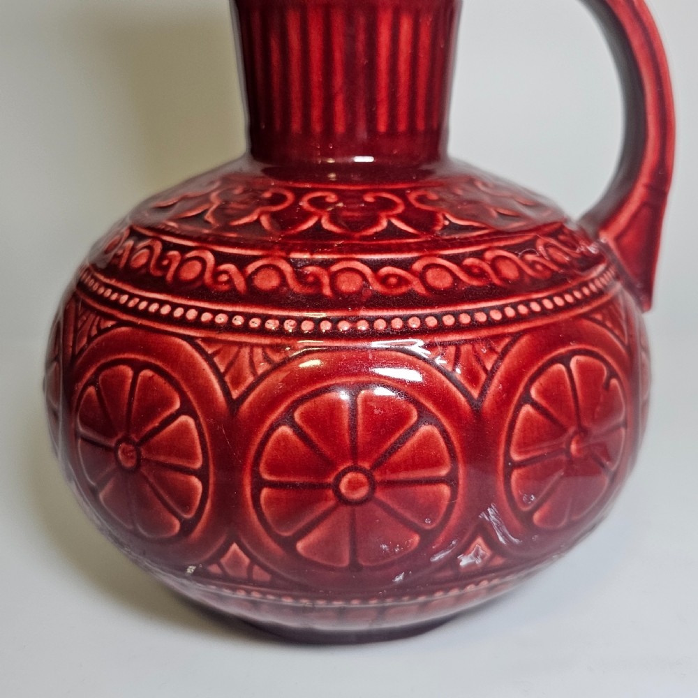 A dark red glazed earthenware jug featuring relief-moulded geometric patterns, a bulbous lower section, a fluted neck, and a curved handle. View VII
