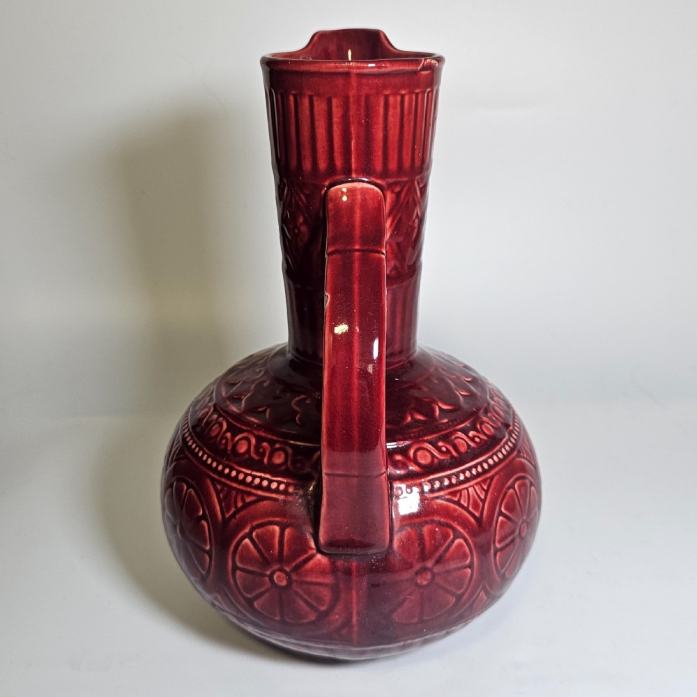 A dark red glazed earthenware jug featuring relief-moulded geometric patterns, a bulbous lower section, a fluted neck, and a curved handle. View VIII