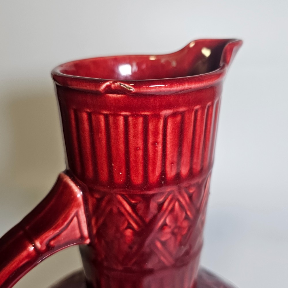 A dark red glazed earthenware jug featuring relief-moulded geometric patterns, a bulbous lower section, a fluted neck, and a curved handle. View IX