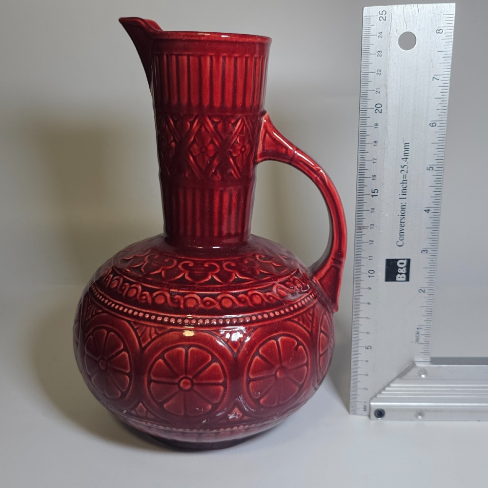 A dark red glazed earthenware jug featuring relief-moulded geometric patterns, a bulbous lower section, a fluted neck, and a curved handle. View X