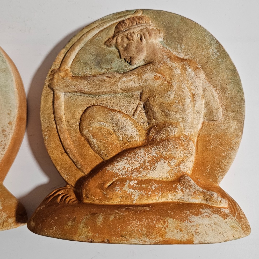 A pair of circular terracotta bookends depicting a kneeling archer with a drawn bow in relief sculpture with a distressed white wash finish. View V