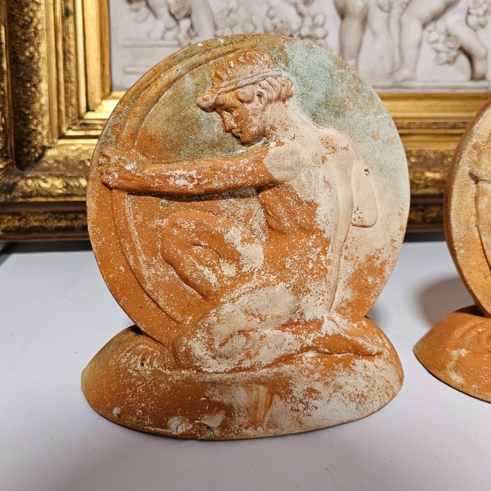 A pair of circular terracotta bookends depicting a kneeling archer with a drawn bow in relief sculpture with a distressed white wash finish. View VI