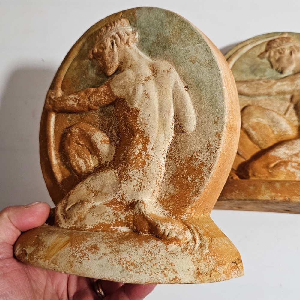 A pair of circular terracotta bookends depicting a kneeling archer with a drawn bow in relief sculpture with a distressed white wash finish. View IX