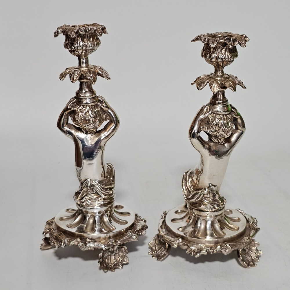A pair of antique silver-plated candlesticks featuring seated cherub figures supporting the candle nozzles above circular scrolled bases. View V