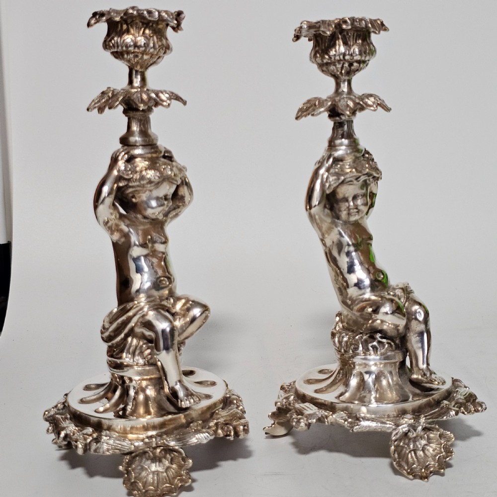 A pair of antique silver-plated candlesticks featuring seated cherub figures supporting the candle nozzles above circular scrolled bases. View II