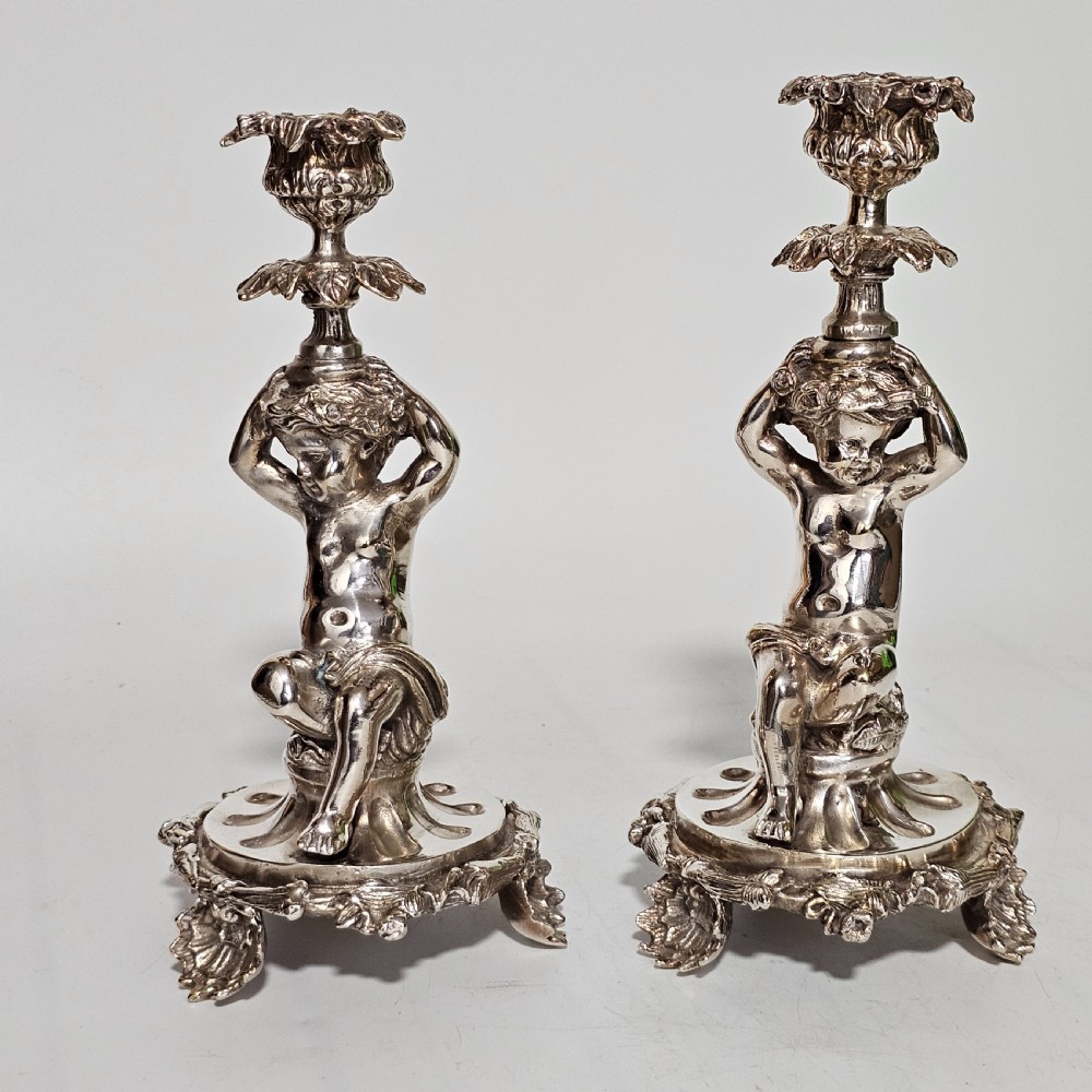 A pair of antique silver-plated candlesticks featuring seated cherub figures supporting the candle nozzles above circular scrolled bases. View III