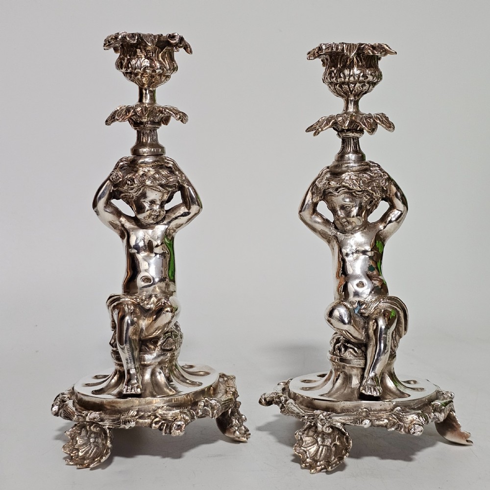 A pair of antique silver-plated candlesticks featuring seated cherub figures supporting the candle nozzles above circular scrolled bases. View IV