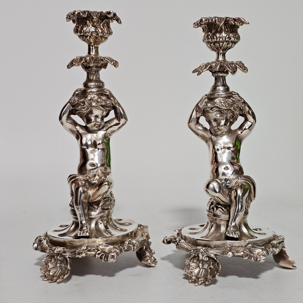 A pair of antique silver-plated candlesticks featuring seated cherub figures supporting the candle nozzles above circular scrolled bases.