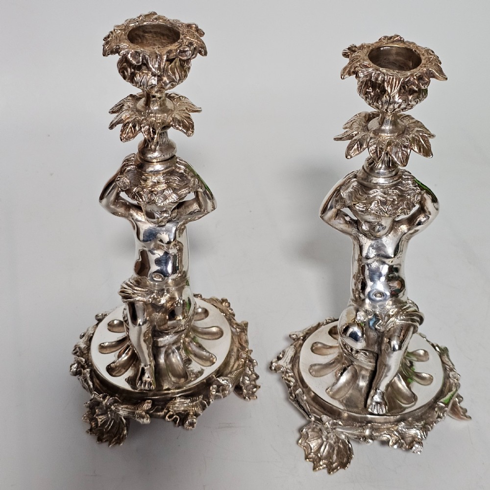 A pair of antique silver-plated candlesticks featuring seated cherub figures supporting the candle nozzles above circular scrolled bases. View VI