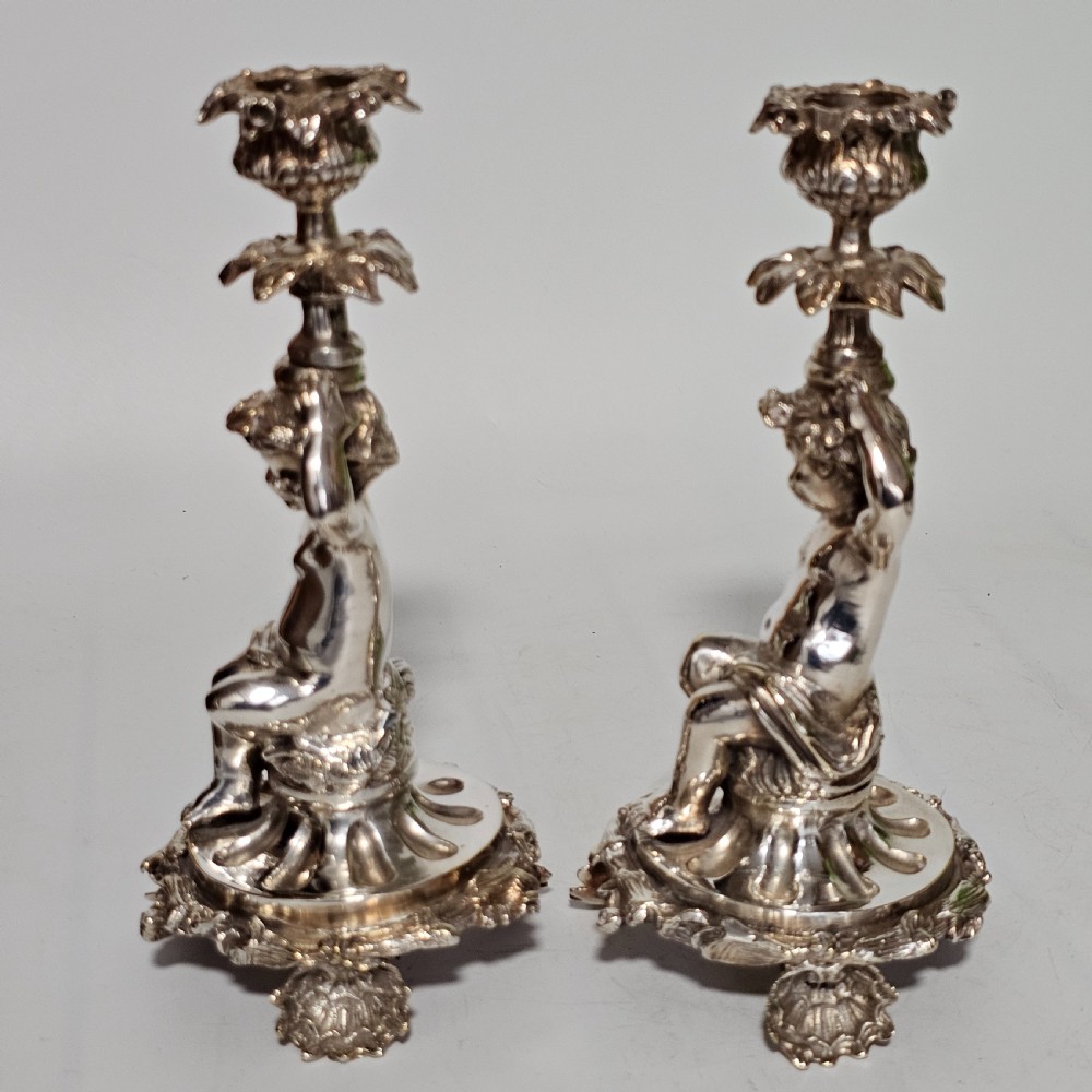 A pair of antique silver-plated candlesticks featuring seated cherub figures supporting the candle nozzles above circular scrolled bases. View VII