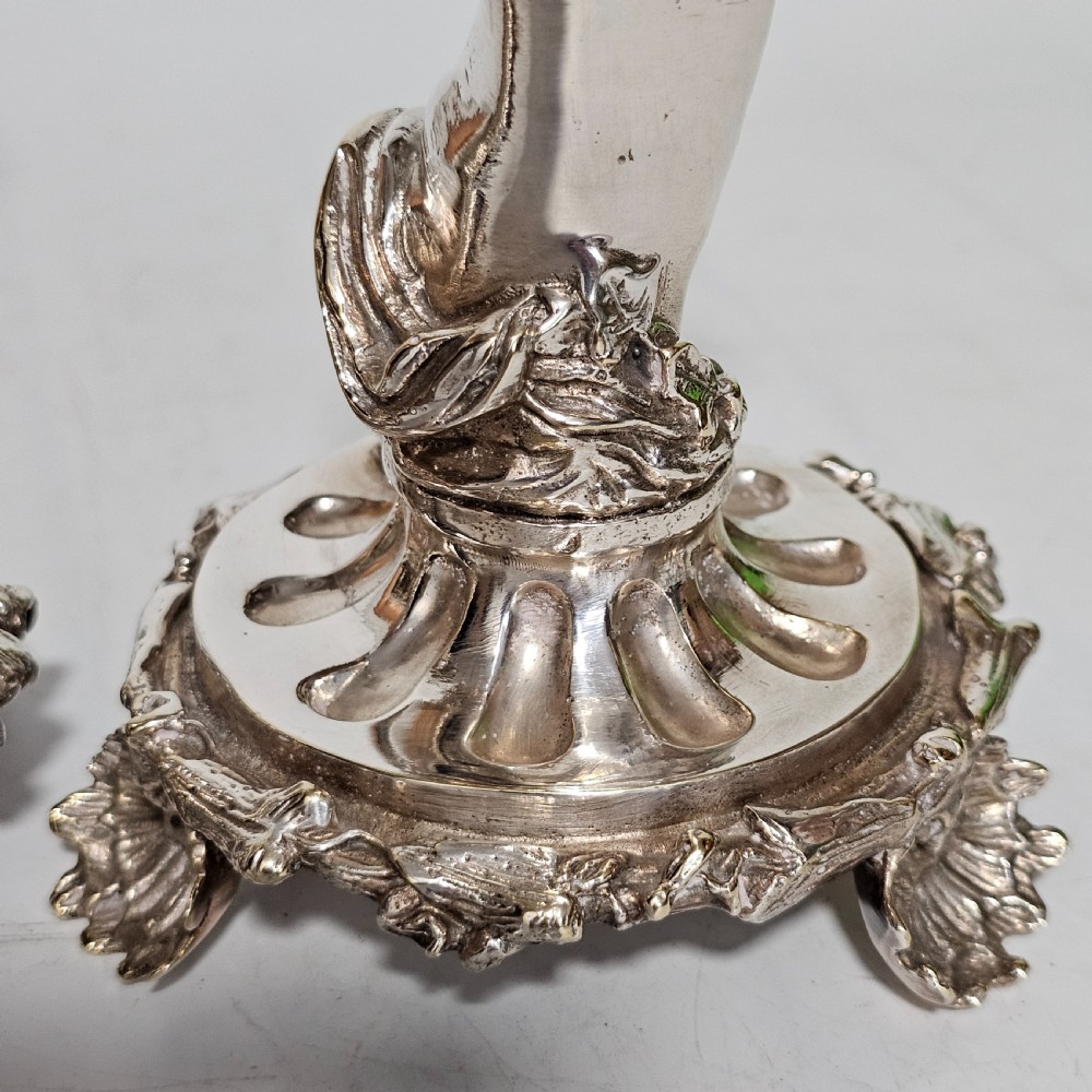 A pair of antique silver-plated candlesticks featuring seated cherub figures supporting the candle nozzles above circular scrolled bases. View IX