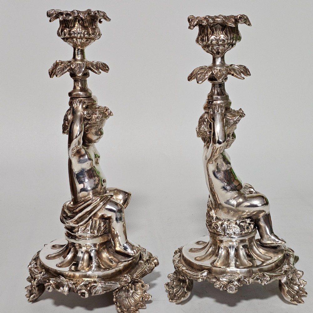 A pair of antique silver-plated candlesticks featuring seated cherub figures supporting the candle nozzles above circular scrolled bases. View X