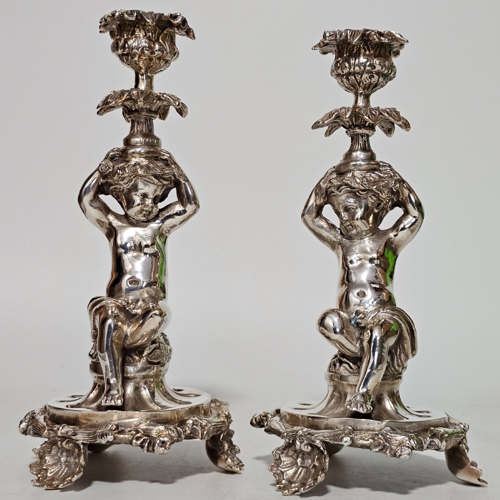 A pair of antique silver-plated candlesticks featuring seated cherub figures supporting the candle nozzles above circular scrolled bases. View XI